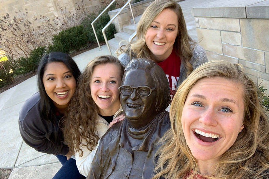A group of four full time MBA students pose for a photo around a statue of Nobel Prize-winner Elinor Ostrom on the Indiana University Bloomington campus.
